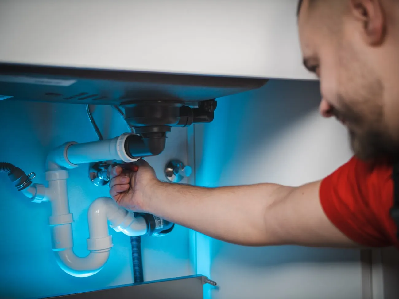 Un plombier règle une évacuation sous un évier, lampe de chantier allumée